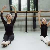 professional asian ballerina with beginner exercising in studio sitting on floor with raised arms