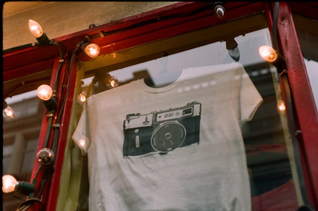 photo of a white t shirt with a camera print on a window display decorated with lights