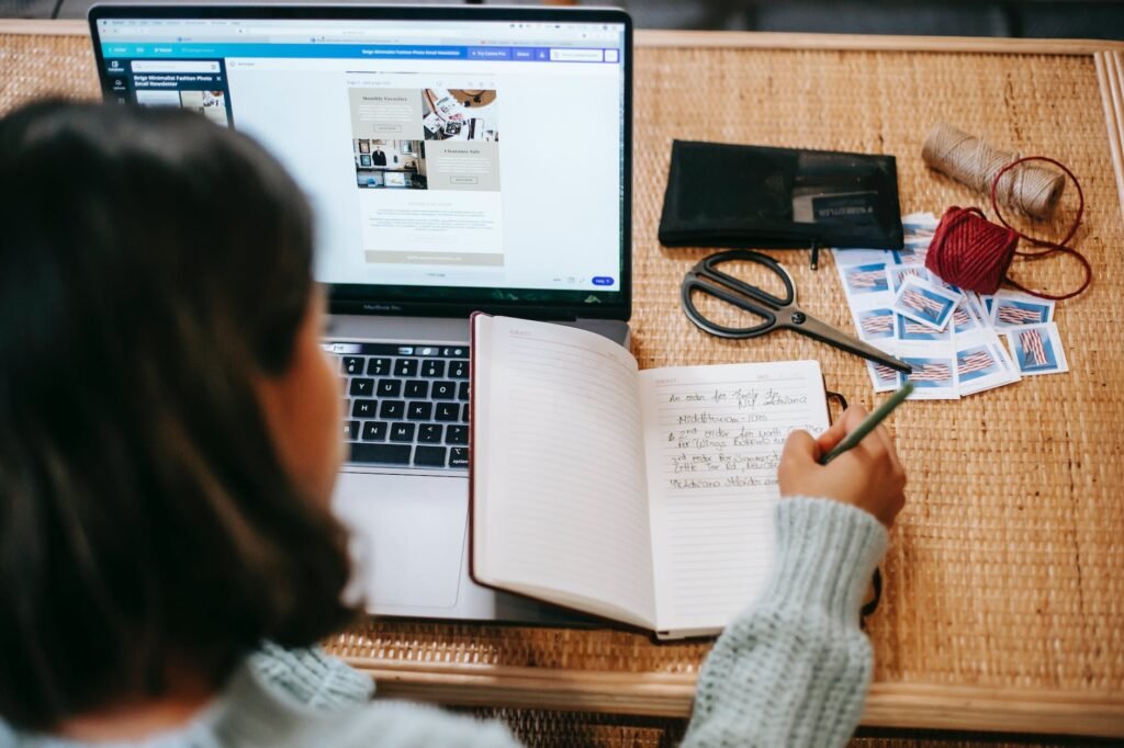 unrecognizable ethnic female student taking notes in notebook near laptop