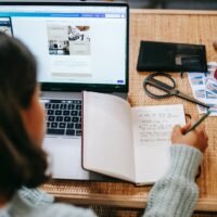 unrecognizable ethnic female student taking notes in notebook near laptop